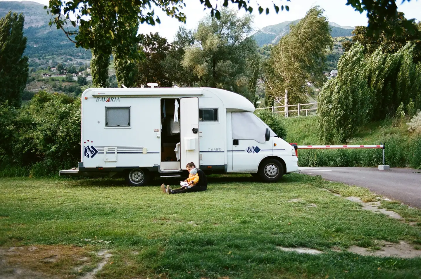 Autocamper på en grøn campingplads med bjerge i baggrunden, forælder og barn sidder i græsset foran